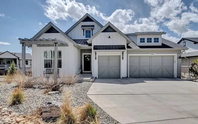 A white modern farmhouse with board and batten siding, grey carriage-style garage doors, and a low-water front yard of rock and native grasses.